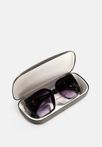 Czarne kwadratowe okulary przeciwsłoneczne w teksturowanym czarnym etui, podszytym miękkim białym wnętrzem. Soczewki mają gradientowy fioletowy kolor, z wygrawerowanym napisem "FRENCH DESIGN" w środku.