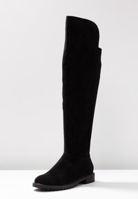 Černé semišové kozačky nad kolena s kulatou špičkou, nízkým blokovým podpatkem a odolnou podrážkou, vystavené na bílé ploše s jednoduchým pozadím.