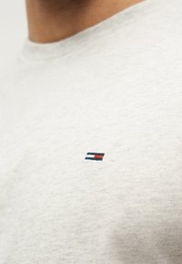 Κοντινό πλάνο ενός ανοιχτό γκρι πουκάμισου με ένα μικρό ορθογώνιο λογότυπο σε ναυτικό μπλε, λευκό και κόκκινο στην περιοχή του στήθους.