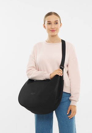 Mladá žena v svetloružovom mikinovom svetri a modrých džínsoch drží veľkú čiernu crossbody tašku Larkson Hamburg na pleci.