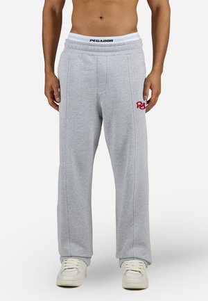 Man wearing light gray sweatpants with red embroidery on thigh and white sneakers, standing against white background.