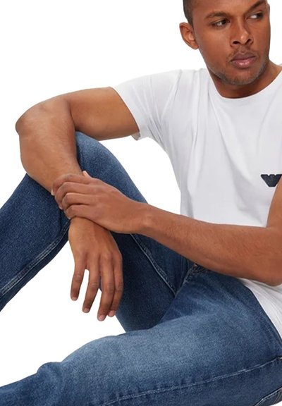 T-shirt blanche à manches courtes avec un logo noir, associée à un jean en denim bleu. Le jean a une coupe slim, avec un motif de couture détaillé.