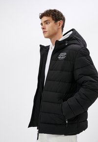 Sort puffers jakke med hætte, med horisontalt kviltet tekstur, lynlåslommer og et logo på venstre brystområde.