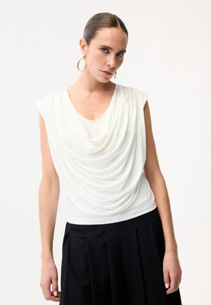 Kobieta ubrana w białą, bezrękawną bluzkę z drapowanym dołem, czarną plisowaną spódnicę i złote kolczyki koła, stojąca na jednolitym jasnym tle.