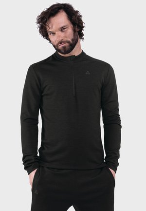 Bärtiger Mann mit lockigem Haar, der ein schwarzes langärmeliges Oberteil mit Reißverschluss und schwarze Hose trägt, mit den Händen in den Taschen vor einem einfarbigen grauen Hintergrund.