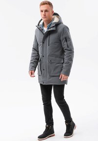 Grå isolerad jacka med huva, fleecefoder, flera fickor och dragkedja fram. Bärs över mörka jeans och svarta stövlar.