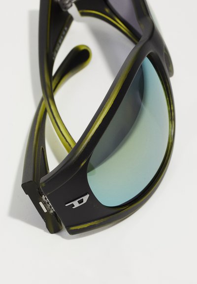 Lunettes de soleil noires avec une monture fine et mate, présentant des accents verts et des verres teintés de bleu. Comprend un détail de logo en argent sur le côté.