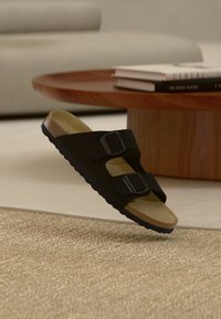 Μαύρα σανδάλια με δύο ρυθμιζόμενες αγκράφες, καφέ φελλό στο πέλμα και υφή μαύρης καουτσούκ σόλας, παρουσιασμένα σε ανυψωμένη θέση.