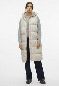Gilet rembourré beige clair avec capuche, doté d'un rembourrage matelassé et d'une fermeture éclair à l'avant. Porté par-dessus un pull gris et un jean sombre.