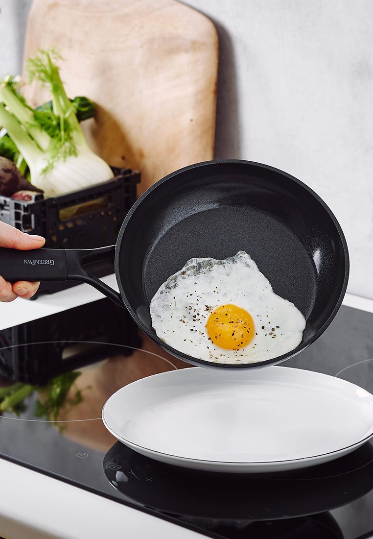 Padella antiaderente nera con un uovo fritto, tenuta sopra un piatto bianco su un piano cottura. Sullo sfondo si vedono un tagliere e delle verdure.