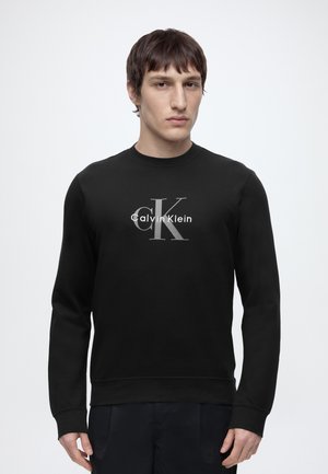 Mand med kort brunt hår iført en sort Calvin Klein sweatshirt med gråt og hvidt logo, stående mod en ensfarvet lys baggrund.