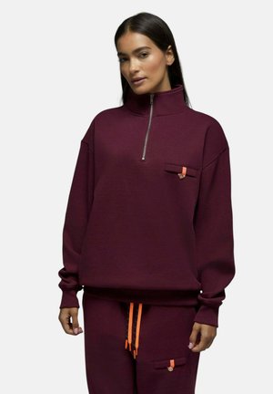 Femme portant un sweat-shirt bordeaux à demi-fermeture éclair et un pantalon assorti avec des cordons orange et des détails de poches boutonnées, debout devant un fond uni.