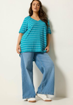 Frau trägt ein türkis gestreiftes V-Ausschnitt-Oberteil, weite hellblaue Jeans und weiße Turnschuhe, steht vor einer einfarbigen hellen Wand.