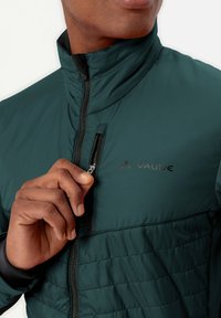 Giacca zip nera con colletto alto, caratterizzata da una texture trapuntata nella parte bassa e da una piccola tasca sul petto. Nome del marchio visibile.
