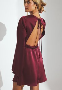Abito di raso bordeaux con maniche lunghe, design con schiena scoperta e lacci, e una vita elasticizzata, che offre una texture liscia e una forma fluida.