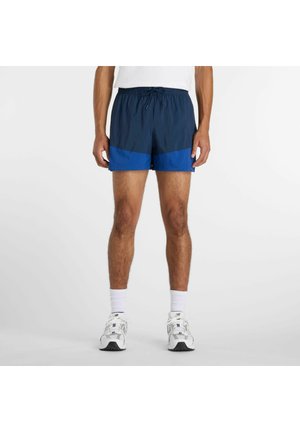 Homme portant un short de sport bleu marine et bleu royal, des chaussettes blanches et des chaussures de course blanches, debout sur un fond clair.