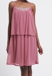 Ärmelloses Kleid in sanftem Rosa, fließendem Chiffonstoff mit einem geschichteten Design und einem verzierten Ausschnitt, der kleine, glänzende Akzente aufweist.