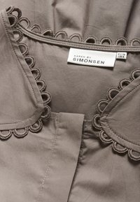Graue Bluse mit gewelltem Ausschnitt, weichem Stoff und einem Etikett, das die Größe EU 38/US 8 angibt. Sauberes und simples Design.