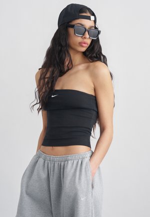 Kobieta w czarnym, bezramkowym topie Nike, szarych spodniach dresowych Nike, czarnych okularach przeciwsłonecznych i czapce odwróconej do tyłu, stoi z rękami w kieszeniach.