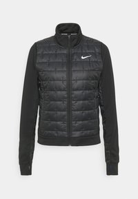 Chaqueta Nike negra con un frontal acolchado, mangas lisas y cuello alto. Cierre de cremallera y logotipo en la parte superior izquierda. Textura suave.