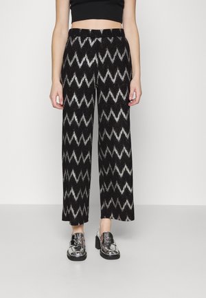 Femme portant un pantalon large à motif zigzag noir et argenté, des mocassins argentés métallisés et un crop top noir.