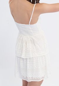 Biała koronkowa sukienka z marszczoną górą, cienkimi regulowanymi ramiączkami, warstwowym, falbanowym wzorem oraz misternymi koronkowymi zdobieniami na całej powierzchni.