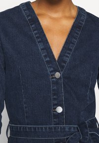 Mörkblå denimklänning med V-ringning, kontrasterande sömmar, fem silvriga knappar och en knytdetalj i midjan. Slät textur.