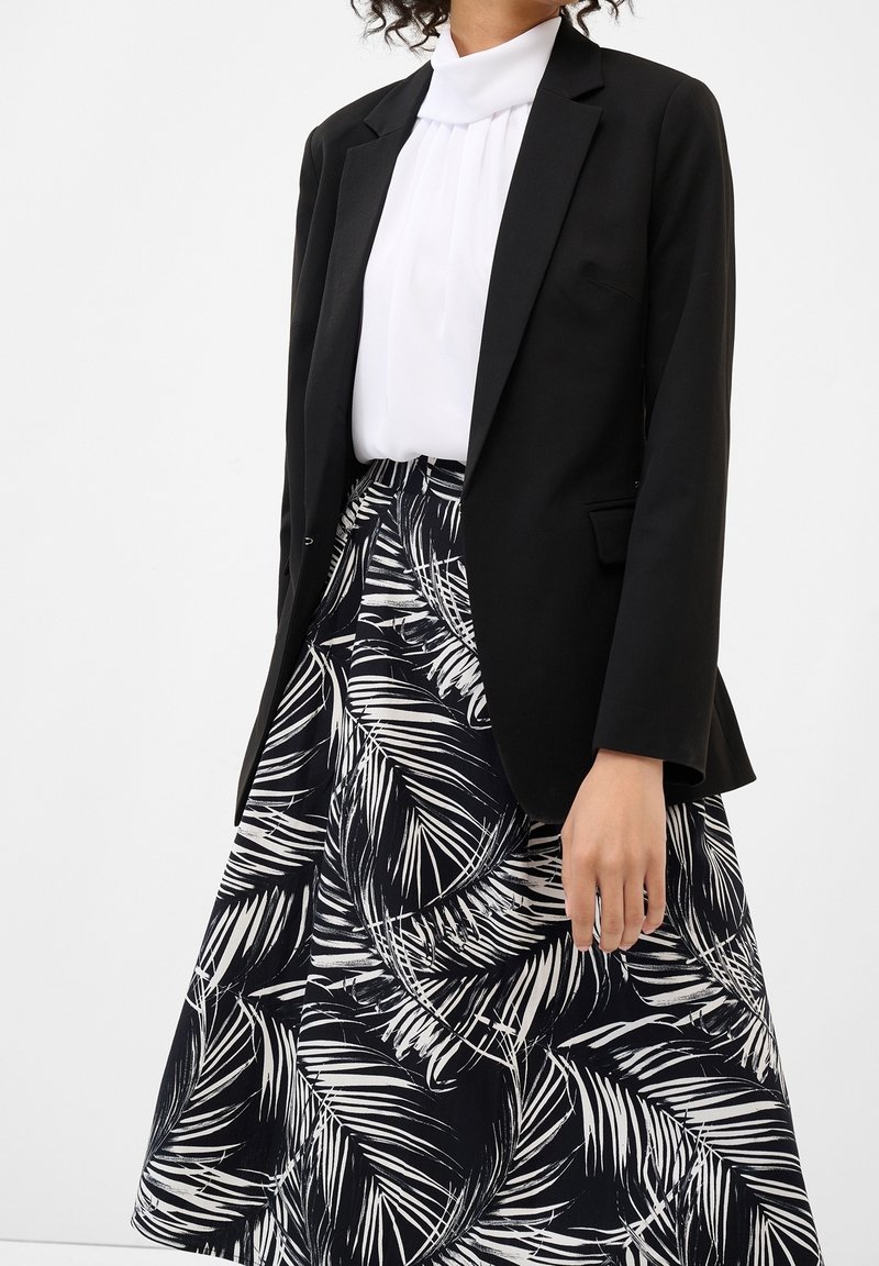 Černý blazer přes bílou plisovanou blůzu, doplněný sukní s černo-bílým vzorem palmových listů. Hladké textury a vypasovaný střih.