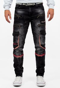 Pantalons en denim noir avec plusieurs poches zippées, accents rouges et coupe fuselée. Présente des coutures visibles et une texture usée.