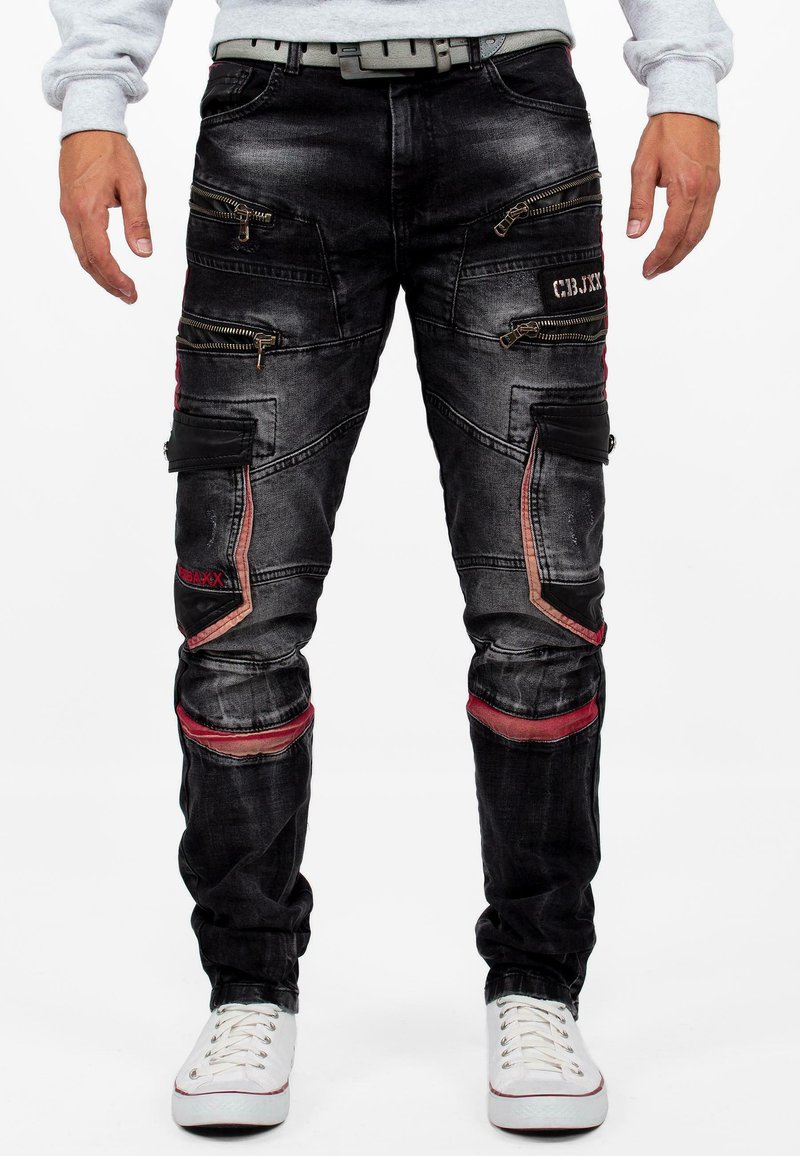 Pantalons en denim noir avec plusieurs poches zippées, accents rouges et coupe fuselée. Présente des coutures visibles et une texture usée.