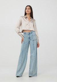 Camicia corta in satin beige chiaro con maniche lunghe e chiusura a bottoni, abbinata a jeans larghi in denim azzurro chiaro con tasche laterali.