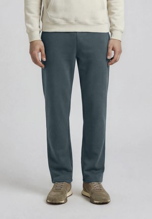 Uomo che indossa una felpa con cappuccio beige, pantaloni dritti grigio scuro e scarpe da ginnastica marrone chiaro, in piedi contro uno sfondo chiaro e semplice.