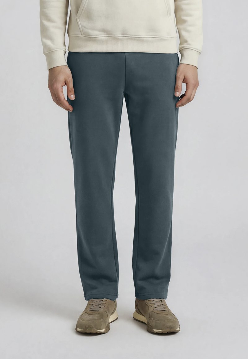 Homme portant un sweat à capuche beige, un pantalon droit gris foncé et des baskets marron clair, debout devant un fond clair uni.