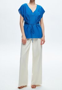Modrá lehká halenka s výstřihem do V, krátkými rukávy a stahovací šňůrkou v pase, kombinovaná s bílými kalhotami s širokými nohavicemi a světlými sandály.