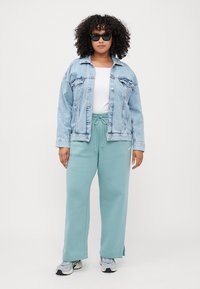 Lys blå denimjakke, hvit t-skjorte og mintgrønne joggebukser med snøring. Modellen har på seg svarte solbriller og sneakers.