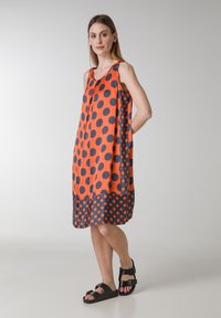 Vestido naranja con lunares grandes negros, diseño sin mangas, forma relajada, dobladillo con picos en la parte inferior, combinado con sandalias negras.