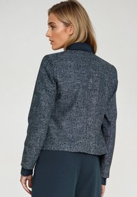 Kobieta zwrócona bokiem, ubrana w teksturowaną szaro-bluzę na czarną koszulę z kołnierzykiem i ciemne spodnie na jednolitym tle.