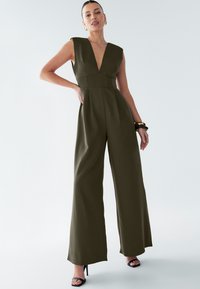 Combinaison olive verte avec décolleté en V profond, design sans manches, silhouette à jambes larges, long torse et détails plissés. Portée avec des sandales à talons.