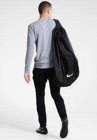 Zwarte Nike sporttas met gaasaccenten, gedragen over de schouder. Grijze lange mouw top, zwarte broek en sportieve schoenen. Eenvoudig, functioneel ontwerp.