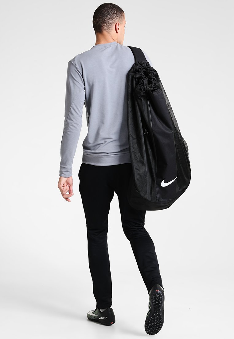 Zwarte Nike sporttas met gaasaccenten, gedragen over de schouder. Grijze lange mouw top, zwarte broek en sportieve schoenen. Eenvoudig, functioneel ontwerp.
