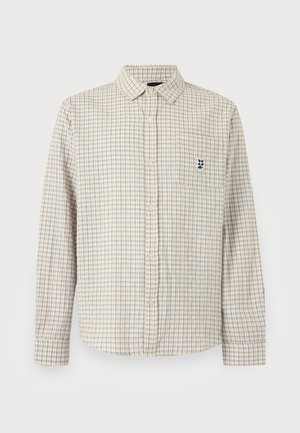 Ung mand iført en beige ternet skjorte med lange ærmer, sorte bukser og en perle halskæde, står mod en hvid baggrund.