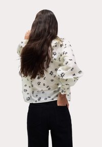 Blusa bianca con motivi floreali neri, maniche a sbuffo e orlo arricciato, abbinata a jeans neri. Tessuto liscio e leggero.