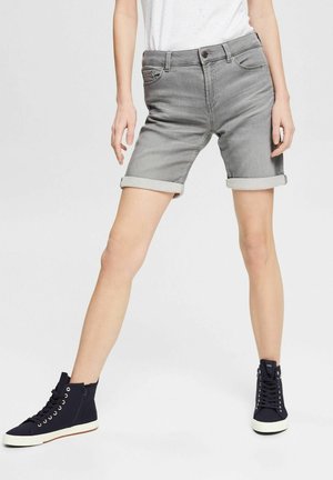 Person iført lysegrå bukser med opkrogede denim shorts, hvid t-shirt og sorte høje sneakers, der står med benene spredt.