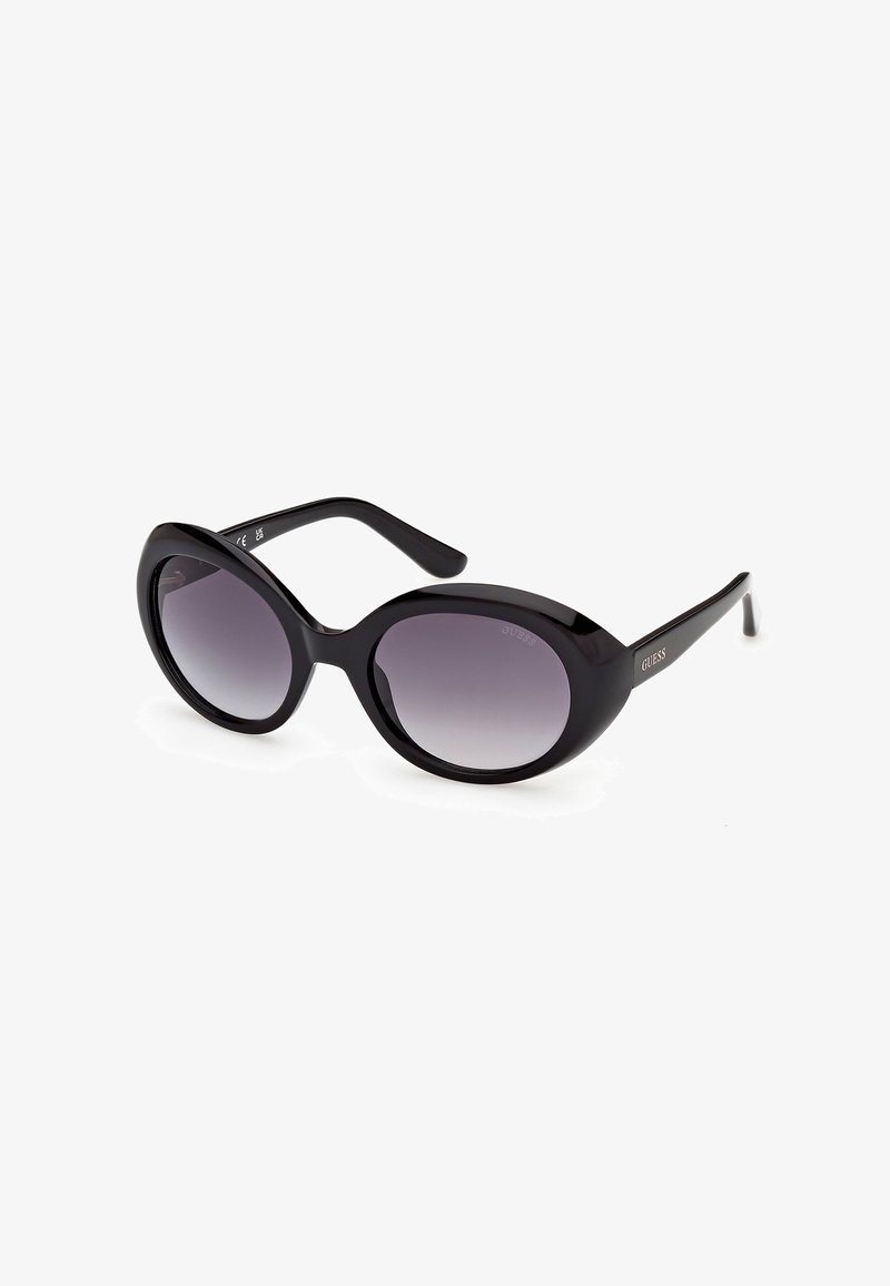 Lunettes de soleil Guess ovales noires avec verres dégradés foncés et montures épaisses et brillantes posées sur un fond blanc.