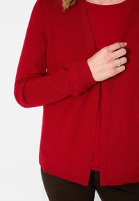 Cardigan rosso lavorato a maglia con apertura anteriore, tessuto strutturato, maniche lunghe e polsini a coste abbinato a una maglietta rossa sottostante.