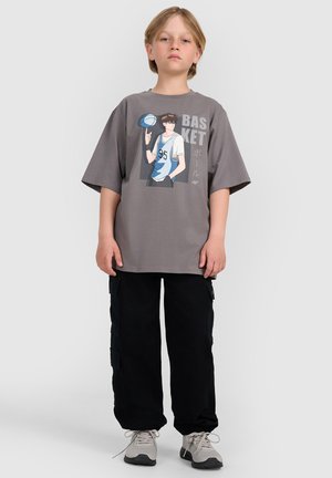 Młoda osoba stoi, ubrana w szarą, oversize koszulkę z grafiką anime o tematyce koszykówki, czarne spodnie bojówki i jasnoszare sneakersy na białym tle.