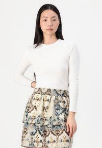 Bluză albă cu mânecă lungă, cu guler rotund, asortată cu o fustă stratificată cu un design patterned în tonuri de bej, albastru și maroniu.