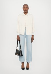 Light beige jacket, relaxed fit with a collar and front pockets. Light blue wide-leg jeans with frayed hems. Black handbag with gold studs.