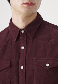 Gros plan d'une personne portant une chemise en velours côtelé bordeaux avec des boutons pression argentés et deux poches poitrine, sur un fond uni.