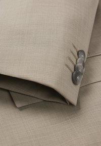 Tessuto beige, finemente strutturato, con un risvolto e tre bottoni grigio scuro, arricchiti da dettagli di cucitura. Due strati visibili sullo sfondo.
